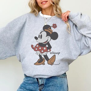 May include: A light gray sweatshirt featuring a Minnie Mouse graphic. The graphic shows Minnie Mouse in a red polka dot skirt, brown shoes, and a flower in her hair. The word "Disney" is printed below the graphic.