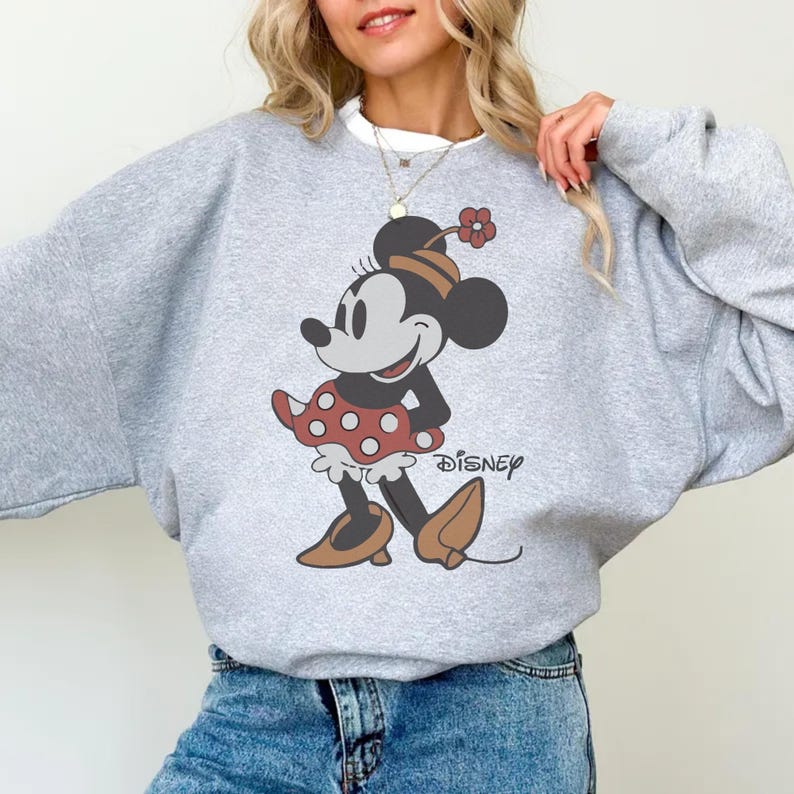 May include: A light gray sweatshirt featuring a Minnie Mouse graphic. The graphic shows Minnie Mouse in a red polka dot skirt, brown shoes, and a flower in her hair. The word "Disney" is printed below the graphic.