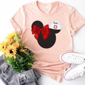 May include: A pink t-shirt with a black and white Mickey Mouse silhouette, a red bow, and a white nurse's hat with a red cross and the name "Lisa" on it.
