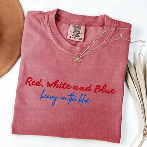 May include: A dusty rose-colored t-shirt with the text "Red, White and Blue heavy on the blue" in red and blue script. A gold pendant necklace and a brown hat are visible. The shirt has a Comfort Colors tag.