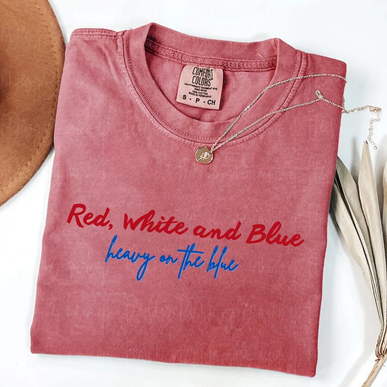 May include: A dusty rose-colored t-shirt with the text "Red, White and Blue heavy on the blue" in red and blue script. A gold pendant necklace and a brown hat are visible. The shirt has a Comfort Colors tag.
