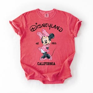 Peut inclure: Un t-shirt rouge avec un dessin de Minnie Mouse portant une robe et un n&oelig;ud &agrave; pois roses. Le t-shirt affiche le logo Disneyland, le texte "est 1955" et "CALIFORNIA" imprim&eacute; en noir.