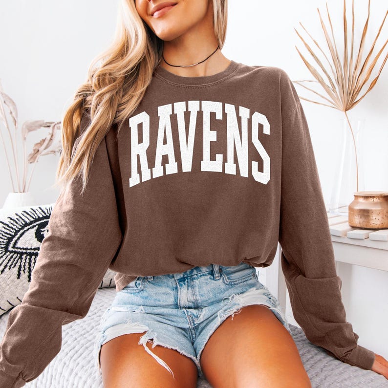 Puede incluir: Una sudadera marr&oacute;n con la palabra "RAVENS" en letras blancas grandes. La parte superior de manga larga se combina con pantalones cortos de mezclilla. La sudadera tiene cuello redondo y un ajuste relajado, adecuada para uso casual.