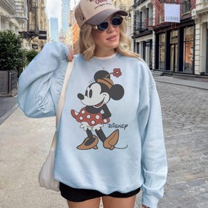 May include: Light blue sweatshirt featuring a Minnie Mouse graphic. The cartoon character is depicted in a red polka dot skirt, brown shoes, and a matching hat. The word "Disney" is printed below the graphic.