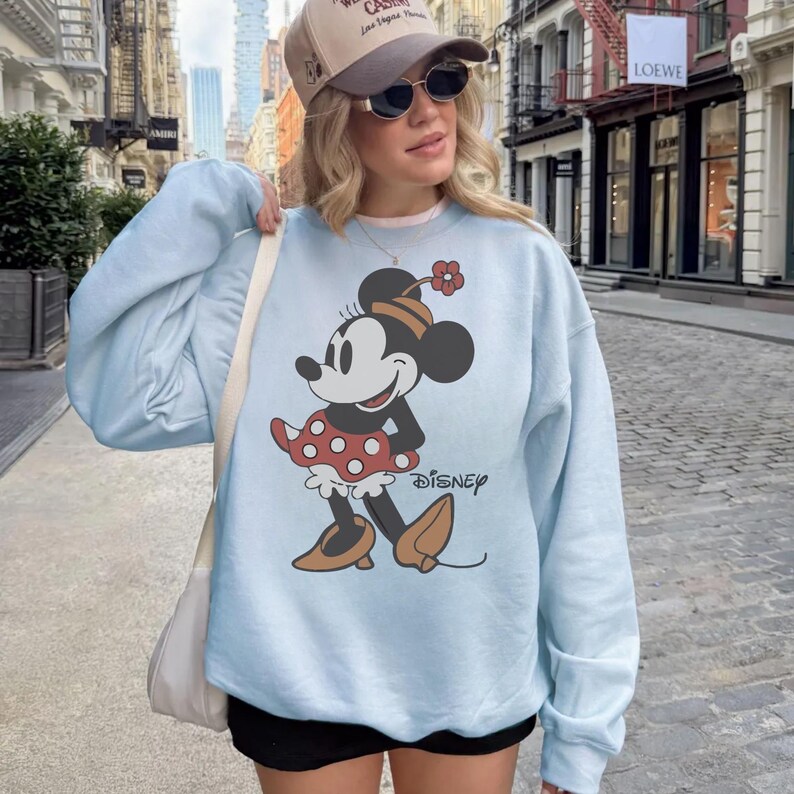 May include: Light blue sweatshirt featuring a Minnie Mouse graphic. The cartoon character is depicted in a red polka dot skirt, brown shoes, and a matching hat. The word "Disney" is printed below the graphic.