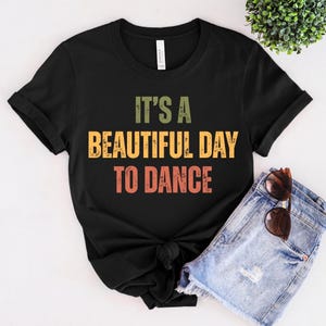 May include: Black t-shirt with the text "IT'S A BEAUTIFUL DAY TO DANCE" in green, yellow, and orange. The shirt is paired with denim shorts and sunglasses. A small green plant is in the background.
