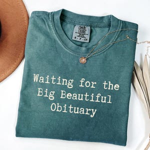 May include: A teal Comfort Colors t-shirt with the text "Waiting for the Big Beautiful Obituary" in white. A gold necklace with a small pendant rests on the shirt. A brown hat is visible in the upper left corner.