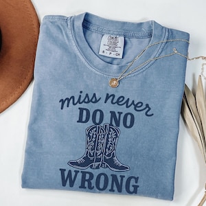 Puede incluir: Una camiseta azul Comfort Colors con el texto "miss never DO NO WRONG" y un gráfico de botas de vaquero. Un collar dorado con un colgante está sobre la camiseta. Un sombrero marrón es visible en la esquina superior izquierda.