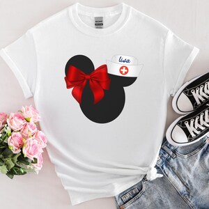 May include: White t-shirt with a black and red Mickey Mouse design. The Mickey Mouse ears have a red bow and a white nurse's hat with a red cross and the name "Lisa" written on it.