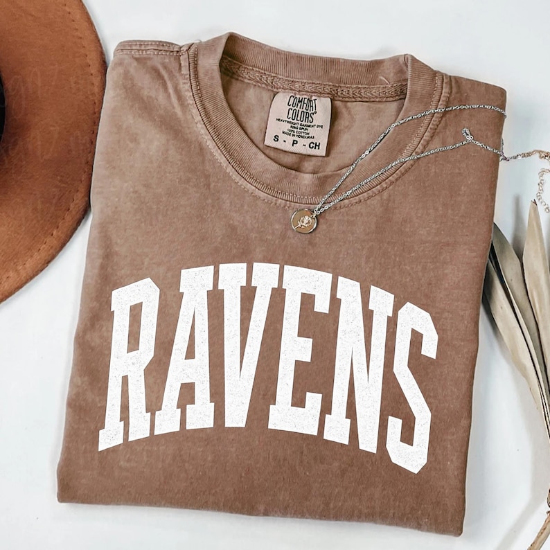 Puede incluir: Una camiseta marr&oacute;n Comfort Colors con la palabra "RAVENS" en letras blancas grandes. Un collar plateado con un peque&ntilde;o colgante descansa sobre la camiseta. La camiseta tiene cuello redondo y mangas cortas.