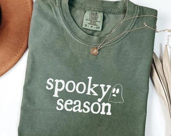 May include: An olive green t-shirt with the words "spooky season" in white, and a ghost graphic. A gold necklace with a pendant rests on the shirt. The shirt is folded, and a brown hat is partially visible in the background.