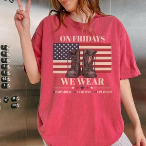 Könnte beinhalten: Rotes T-Shirt mit dem Text "ON FRIDAYS WE WEAR" über einer amerikanischen Flaggen-Grafik und zwei Kampfstiefeln. Der Text "REMEMBER EVERYONE DEPLOYED" befindet sich darunter. Das Shirt ist einfarbig.