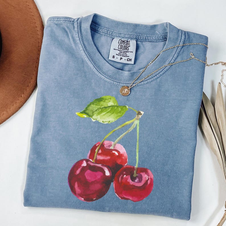 May include: A light blue t-shirt featuring a watercolour painting of three red cherries with green leaves. The shirt has the text "COMFORT COLORS" and "S - P - CH" printed on the tag.