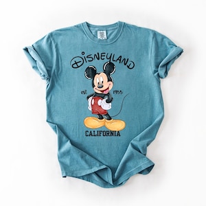 Peut inclure: T-shirt bleu sarcelle avec un dessin de Mickey Mouse. Le texte "Disneyland" est au-dessus et "California" en dessous. Mickey porte un short rouge, des gants blancs et des chaussures jaunes. Les manches sont retrouss&eacute;es.