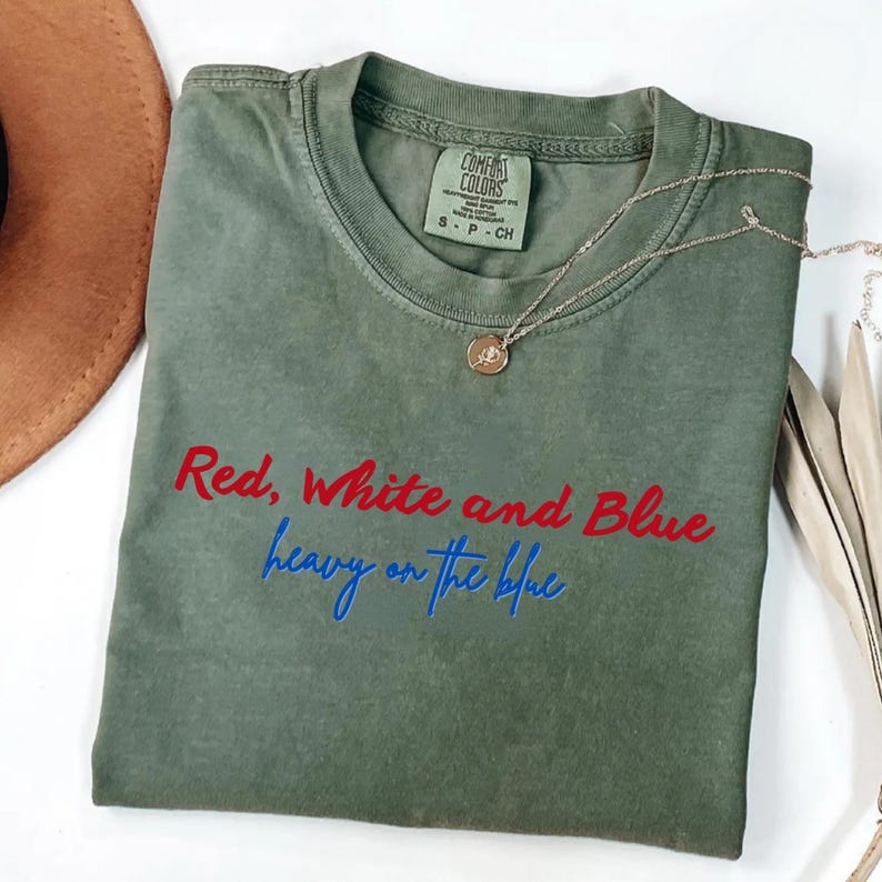 May include: An olive green t-shirt with the text "Red, White and Blue heavy on the blue" in red and blue lettering. A gold necklace with a pendant is draped across the shirt. The shirt is a Comfort Colors brand.