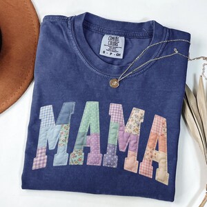 Op de afbeelding: Een marineblauw Comfort Colors T-shirt met het woord "MAMA" in patchworkstof. De patchworkletters hebben verschillende bloemen- en patroonontwerpen. Een gouden ketting met een hanger ligt op het shirt.
