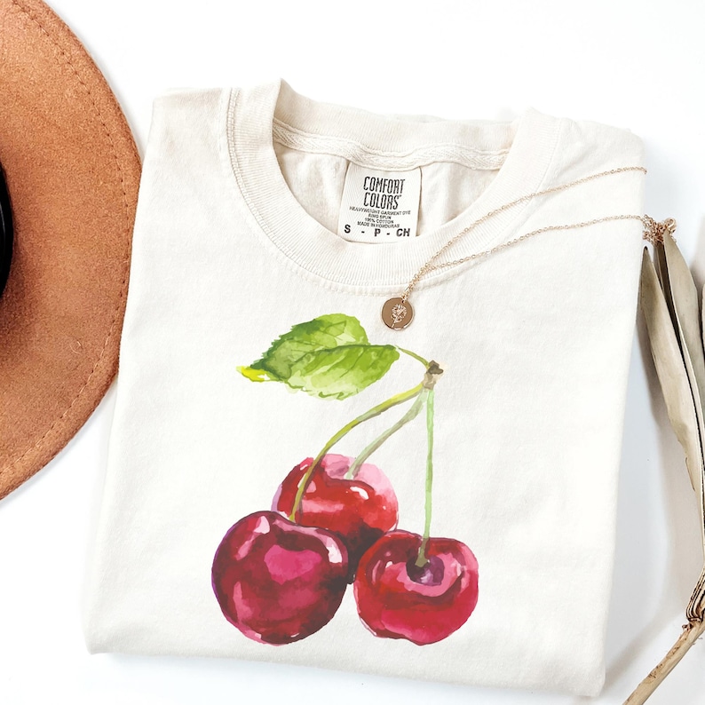 May include: A white t-shirt featuring a watercolour illustration of three red cherries with green leaves. The shirt has a tag that reads "Comfort Colors" on it.
