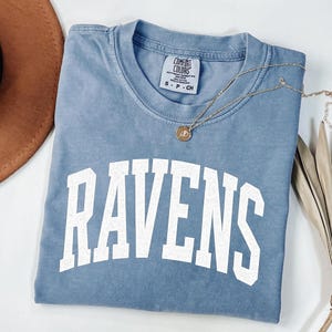 Puede incluir: Una camiseta azul Comfort Colors con la palabra "RAVENS" en letras blancas grandes. Un collar de oro con un colgante descansa sobre la camisa. La camisa est&aacute; doblada y se muestra con un sombrero marr&oacute;n y tallos de plantas secas.