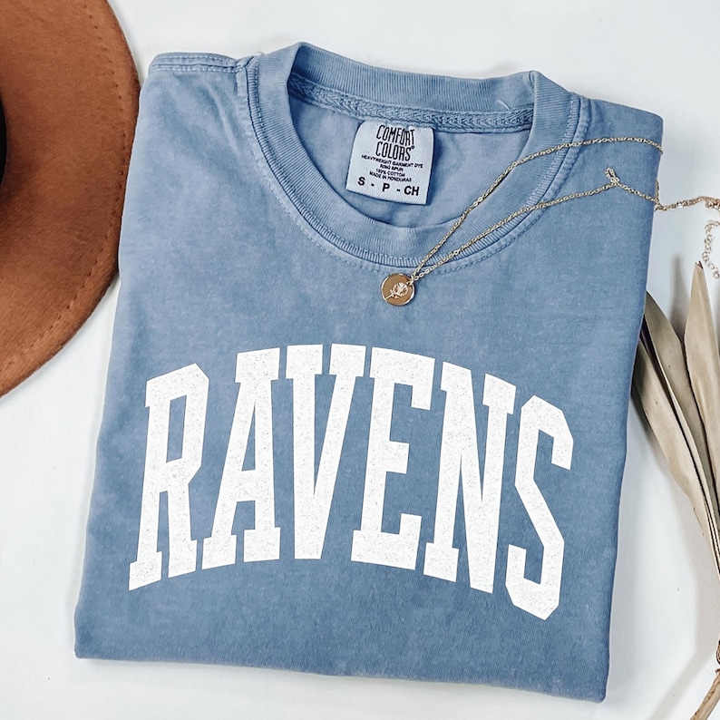 Puede incluir: Una camiseta azul Comfort Colors con la palabra "RAVENS" en letras blancas grandes. Un collar de oro con un colgante descansa sobre la camisa. La camisa est&aacute; doblada y se muestra con un sombrero marr&oacute;n y tallos de plantas secas.