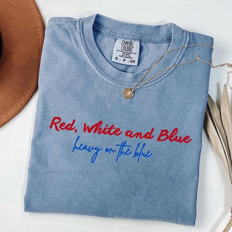 May include: A light blue Comfort Colors t-shirt with the text "Red, White and Blue heavy on the blue" in red and blue. A gold necklace with a pendant is draped across the shirt. A brown hat is visible in the background.