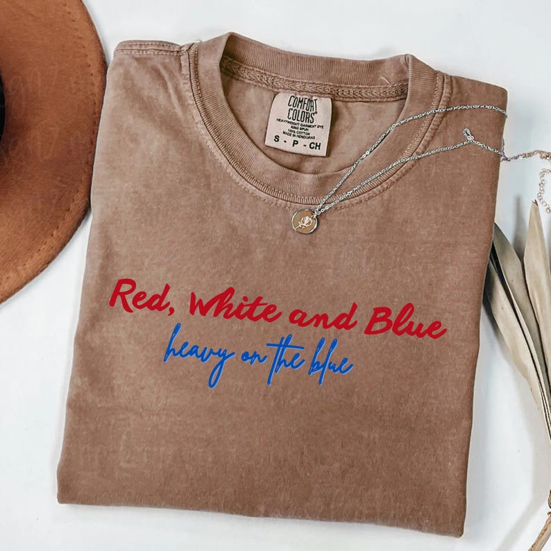 May include: A brown Comfort Colors t-shirt with the text "Red, White and Blue heavy on the blue" in red and blue. A silver necklace with a small pendant is draped across the shirt.