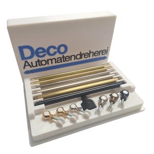 Könnte beinhalten: Eine weiße Box mit verschiedenen Metallstäben und dekorativen Schrauben. Die Stäbe sind in Gold, Silber und Schwarz gehalten. Auf der Box steht in blauer Schrift "Deco Automatendreherei".