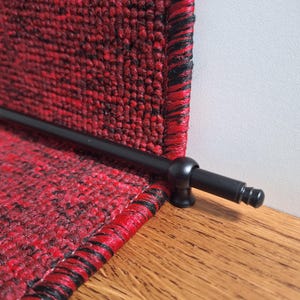May include: Close-up of a black metal stair rod with a rounded end. The rod is attached to a red and black patterned carpet runner.