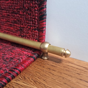 Könnte beinhalten: Eine goldene Treppenstange aus Metall mit abgerundetem Endstück. Die Stange ist an einem roten und schwarzen gemusterten Teppich befestigt.