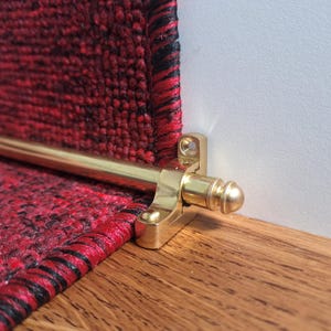 May include: Close-up of a gold metal stair rod with a round finial. The rod is mounted to a wall and a red carpet is visible in the background.