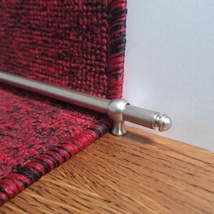 May include: A silver metal stair rod with a rounded end cap. The rod is attached to a red carpet with black edging.
