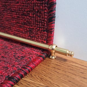 May include: Close-up of a stair runner with a brass rod. The runner is a deep red with black accents, and the brass rod is attached to the wooden step. The runner is made of a woven material.