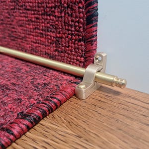 Könnte beinhalten: Messingtreppenstange mit abgerundetem Ende und einer Halterung zur Befestigung an einer Treppenstufe. Die Stange ist vor einem roten und schwarzen Teppich gezeigt.