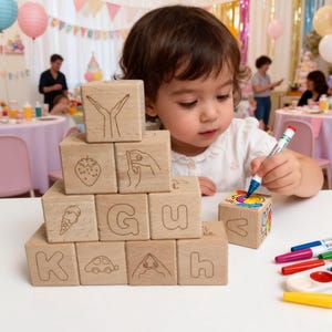 May include: Wooden alphabet blocks with engraved images and letters, stacked in a pyramid shape. A child is using a marker to colour a block. The blocks feature images of a strawberry, ice cream cone, and a car.