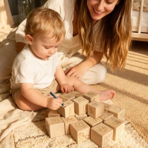 Peut inclure: Un enfant dessine sur des blocs d'alphabet en bois. Les blocs sont de couleur claire avec des illustrations noires de lettres et de symboles. L'enfant est assis sur un tapis à motifs avec une femme en arrière-plan.