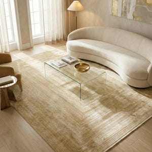 May include: A contemporary living room scene with a beige patterned rug, a curved cream sofa, and a glass coffee table. The room is illuminated by natural light from a large window, creating a warm and inviting atmosphere. The rug's pattern is geometric.