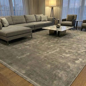 May include: A contemporary living room with a large grey corner sofa, two armchairs, and a marble-topped coffee table. A large grey rug covers the floor, and sheer curtains frame the windows. A floor lamp provides soft lighting.