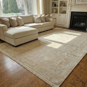 May include: A large, rectangular beige rug with a fringed edge, placed on a hardwood floor. A beige sectional sofa with several cushions is in the background. The room has a fireplace and large windows.