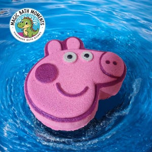 May include: A pink bath bomb in the shape of a cartoon pig's head, with purple detailing, floating in blue water. The image includes a circular logo with a green cartoon dinosaur and the text "MAGIC BATH MOMENTS HANDMADE BATH BOMBS."