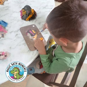 May include: A child decorates a heart-shaped bath bomb on a metal tray. The bath bomb is white with black text and orange accents. Other colourful bath bombs are on the marble surface. The image includes a logo that says "Magic Bath Moments, Handmade Bath Bombs."