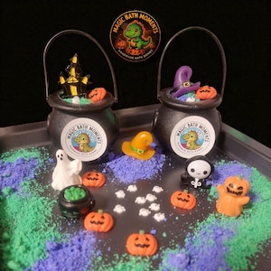 May include: A Halloween-themed arrangement with two black cauldron bath bombs, each with miniature decorations. The scene includes orange pumpkins, a ghost, a witch's hat, and the text "MAGIC BATH MOMENTS HANDMADE BATH BOMBS". Green and purple powder surrounds the items.