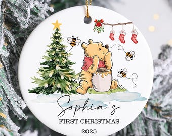 May include: A white ceramic Christmas ornament featuring Winnie the Pooh, a Christmas tree, and bees. The ornament has the text "Sophia's FIRST CHRISTMAS 2025". The ornament is hanging on a Christmas tree.