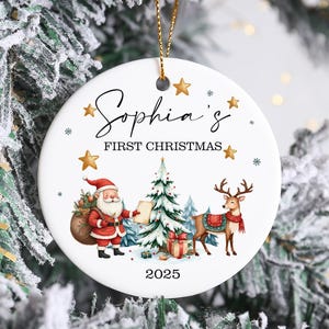 May include: A white ceramic Christmas bauble with a gold string, featuring the text "Sophia's First Christmas" and festive illustrations of Santa Claus, a Christmas tree, and a reindeer. The year 2025 is also displayed.