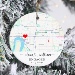 May include: A white ceramic Christmas ornament with a map design, a red heart, and the names "elena & william" with the word "ENGAGED" and the date "5.18.2025". The ornament hangs from a gold string and is surrounded by a snowy Christmas tree.