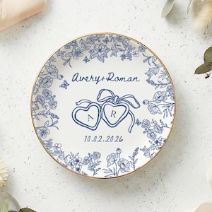 May include: A white ceramic plate with a gold rim, featuring a blue floral design and the names "Avery + Roman" with the date "10.02.2026". Two heart-shaped designs with the letters "A" and "R" are also present. The plate is surrounded by flowers.