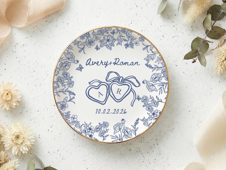 May include: A white ceramic plate with a gold rim, featuring a blue floral design and the names "Avery + Roman" with the date "10.02.2026". Two heart-shaped designs with the letters "A" and "R" are also present. The plate is surrounded by flowers.