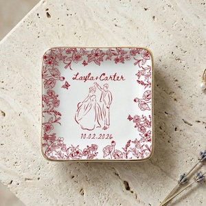 May include: Square white ceramic ring dish with a red floral border and a red illustration of a couple dancing. The dish has gold trim and the names "Layla + Carter" and the date "10.02.2026" are printed on the dish.