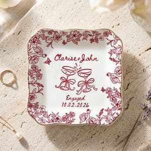 May include: A white square trinket dish with a red floral border and gold trim. The dish features the names "Clarise + John", two champagne glasses, and the date 10.02.2026, with the word "Engaged" below the glasses.
