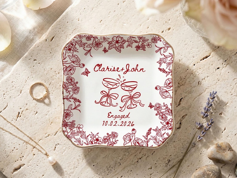 May include: A white square trinket dish with a red floral border and gold trim. The dish features the names "Clarise + John", two champagne glasses, and the date 10.02.2026, with the word "Engaged" below the glasses.
