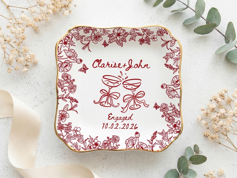 May include: Square white ceramic plate with a gold rim, adorned with red floral patterns and the words "Clarise + John" and "Engaged 10.02.2026". Two champagne glasses are depicted in the center.