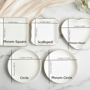 May include: A collection of white ceramic plates in various shapes: square, scalloped, heart, and two circles. Each plate has a gold rim and measurements in inches and centimeters. The square plate is 3.54 inches (9cm) and the heart plate is 3.94 inches (10cm).